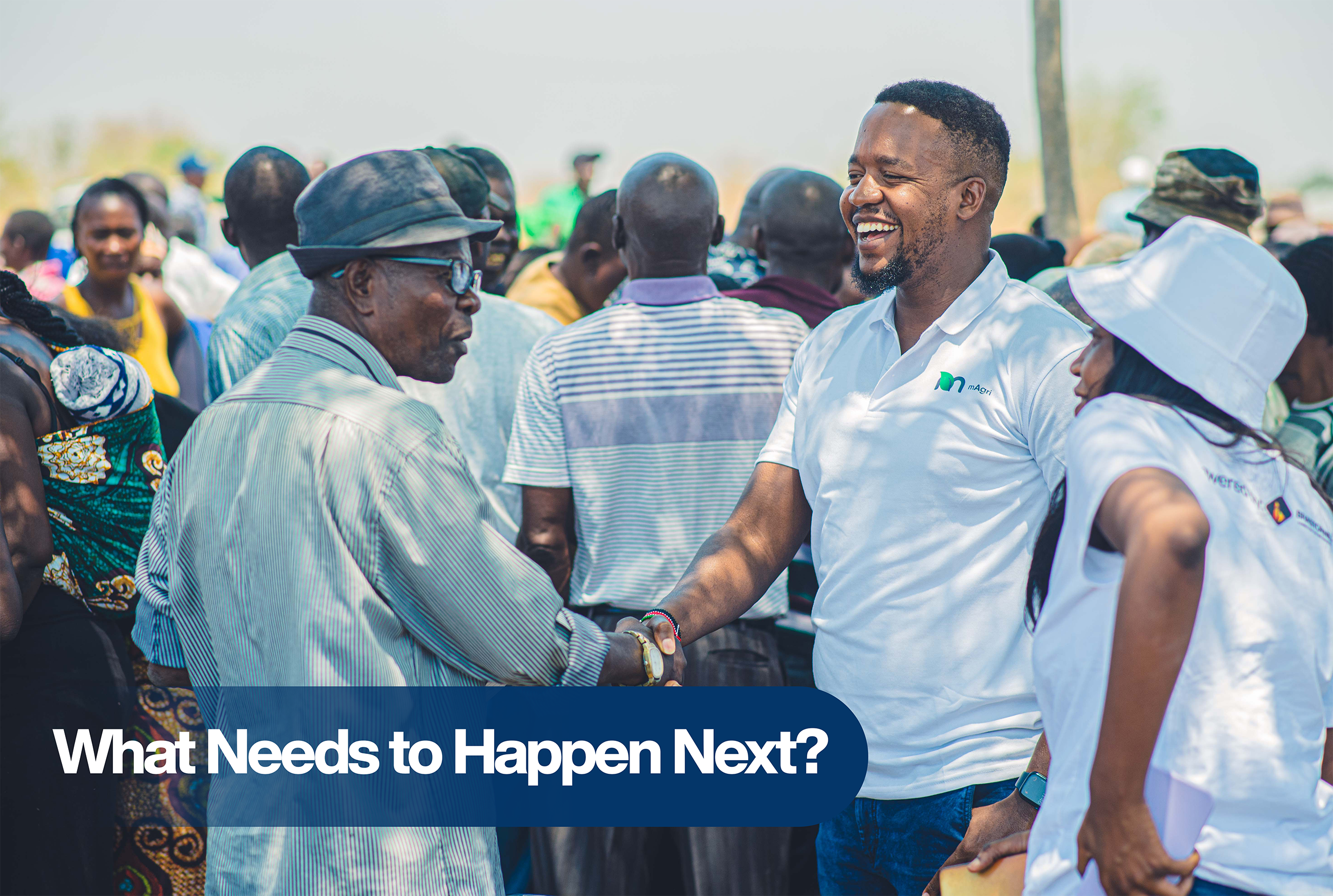 A smiling man in a white mAgri-branded shirt warmly shakes hands with an elderly man at a community gathering, surrounded by farmers and local residents. The atmosphere is vibrant and welcoming, reflecting a sense of unity and shared purpose. Text overlay reads: “What Needs to Happen Next?”